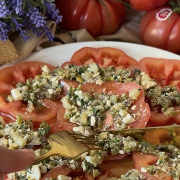 Ensalada de tomate con mezcla de queso feta, pepinillos, alcaparras, cebolleta, anchoas, aceitunas y albahaca.
