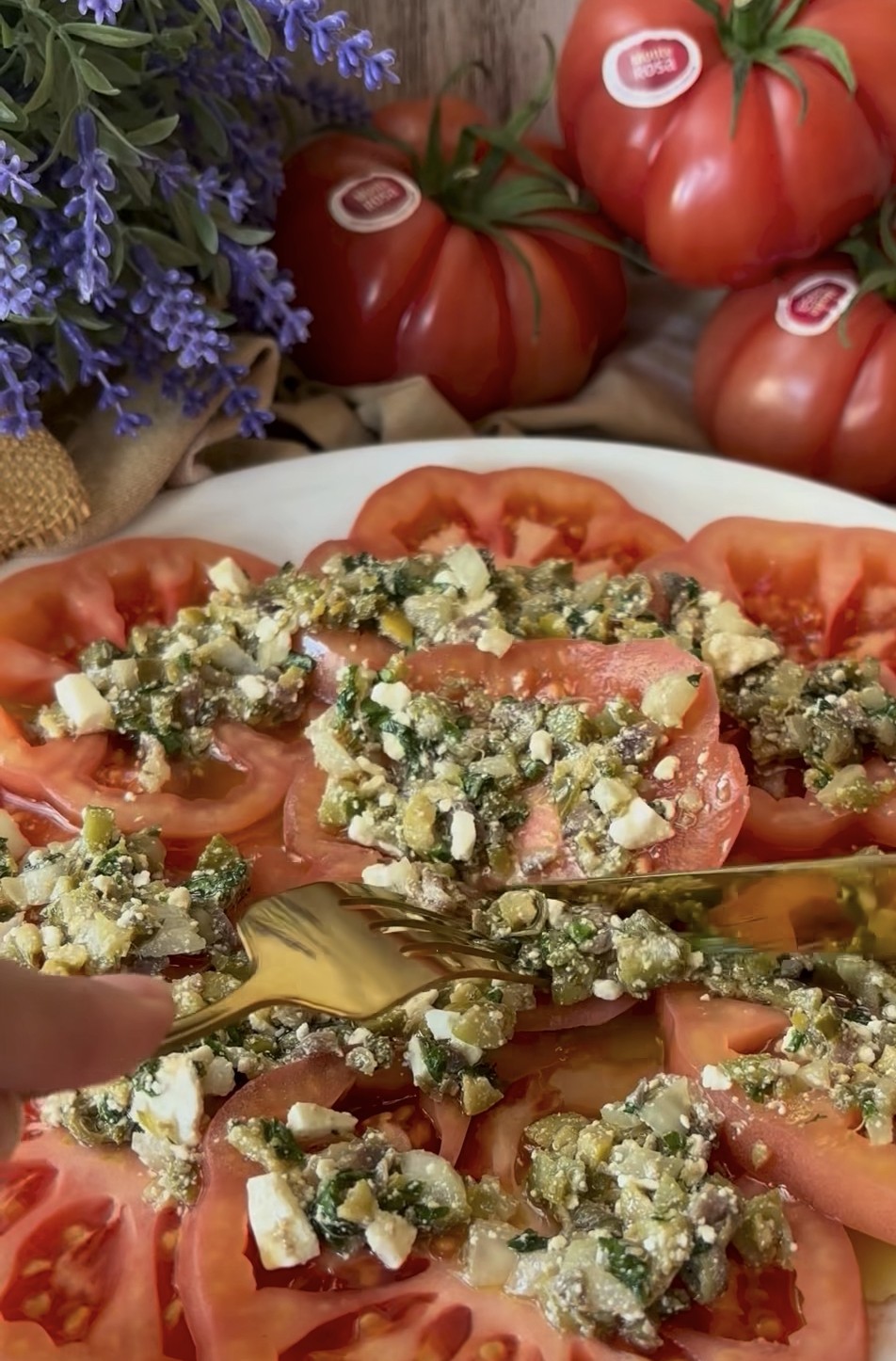 Ensalada de tomate con mezcla de queso feta, pepinillos, alcaparras, cebolleta, anchoas, aceitunas y albahaca.